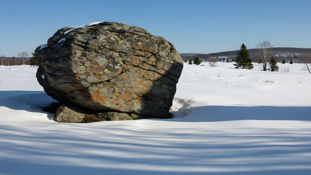 Großer Fels im verschneiten Feld unter blauem Himmel.